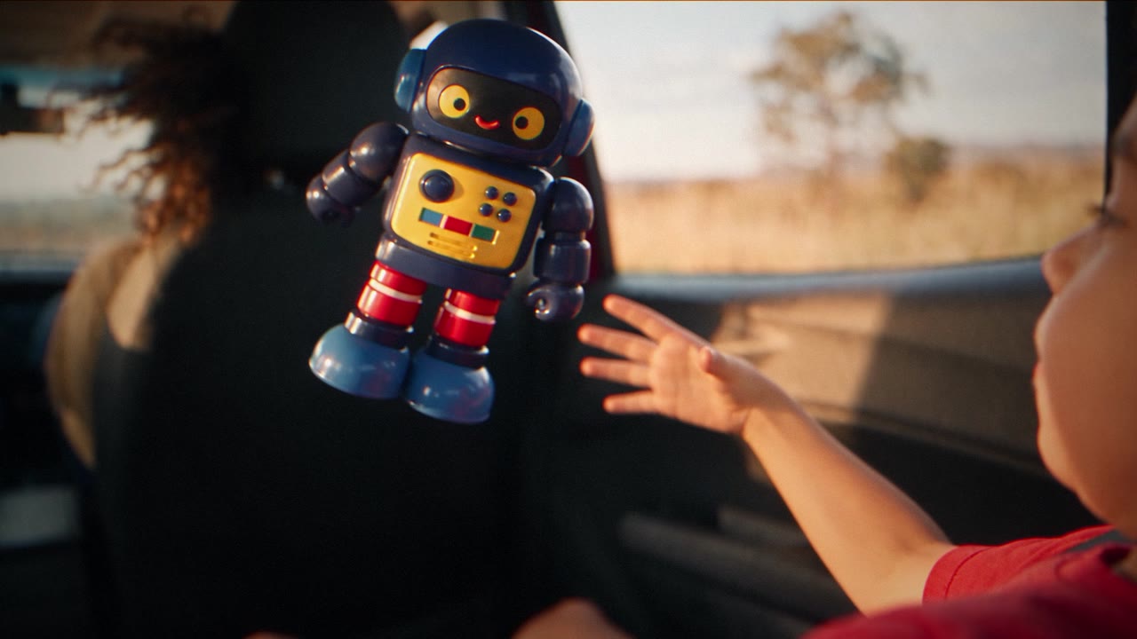 Robô de brinquedo azul no ar diante de uma criança dentro de um carro, em cena de Mostramed — Acidente.